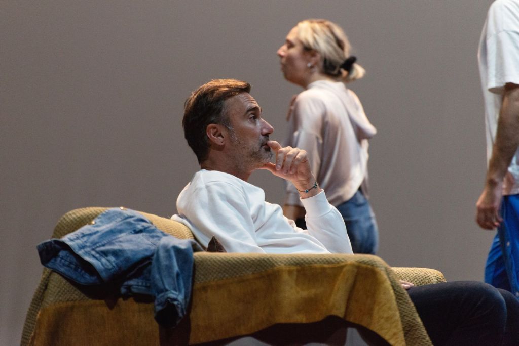 Ο Γιώργος Καπουτζίδης καθισμένος σε μια πολυθρόνα παρακολουθεί τους ηθοποιούς του 