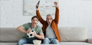 Νεαρος μαζί με ώριμο άνδρα πανηγυριζουν αγώνα ποδοσφαίρου που βλέπουν στην τηλεόραση