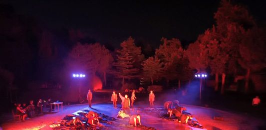 Ηθοποιοί σε κόκκινο και μπλε φωτισμό πάνω σε κυκλική υπαίθρια σκηνή με δέντρα στο φόντο, στιγμιότυπο από την παράσταση Άη Λαός στο Θέατρο Γης